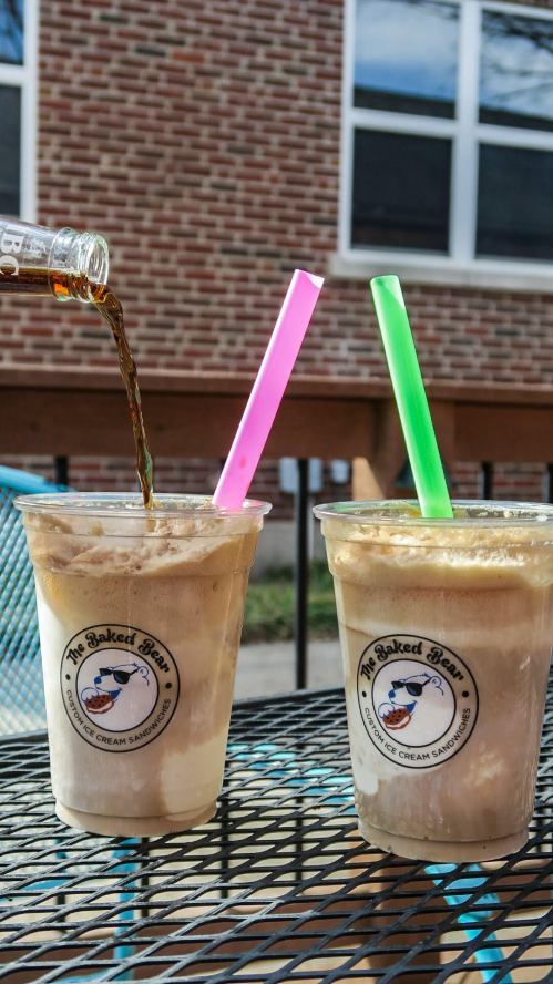 Two ice cream floats in clear cups with colorful straws, one being poured with soda, on a table outdoors.