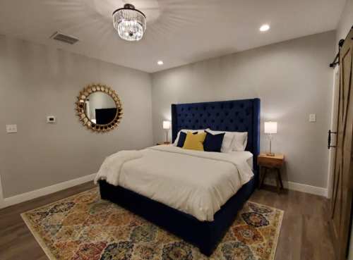 A cozy bedroom featuring a blue tufted bed, decorative mirror, and warm lighting on wooden nightstands.