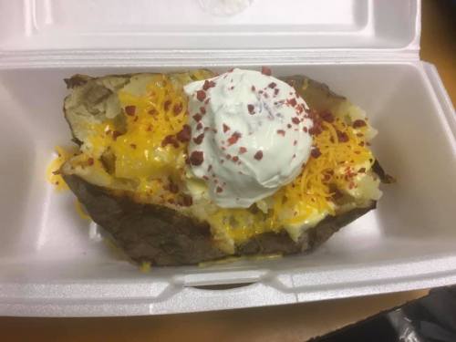 A loaded baked potato topped with cheese, sour cream, and bacon bits in a takeout container.