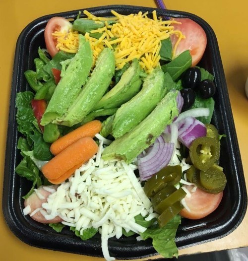 A colorful salad with lettuce, tomatoes, avocado, cheese, olives, carrots, onions, and jalapeños in a black container.