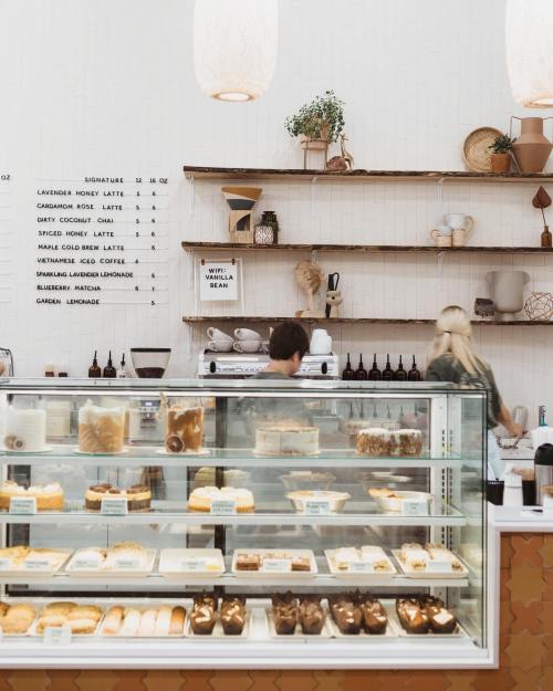 A cozy café interior with a display case of pastries and a menu on the wall, featuring two baristas at work.