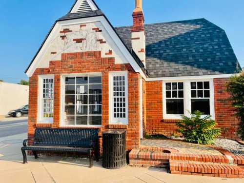 A charming brick building with a sloped roof, large windows, and a bench in front, set against a clear blue sky.