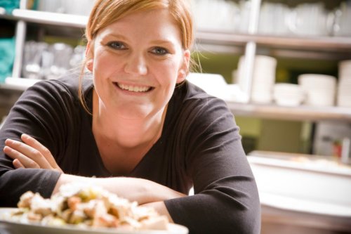 A smiling woman in a black shirt leans on a counter, showcasing a plate of food in a restaurant kitchen.
