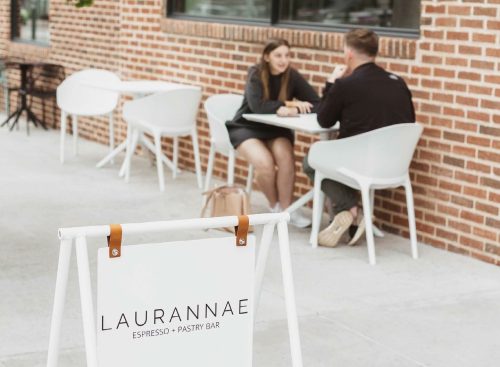 A sign for Laurannae Espresso & Pastry Bar in front of a café where two people are seated at a table.