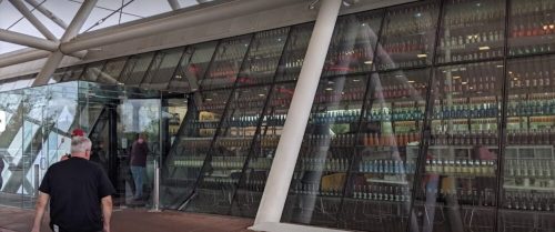 A modern building with large glass windows displaying colorful bottles inside, with a person walking by.
