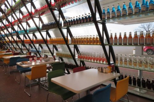 A colorful display of soda bottles lines a modern café wall, with empty tables and chairs in the foreground.