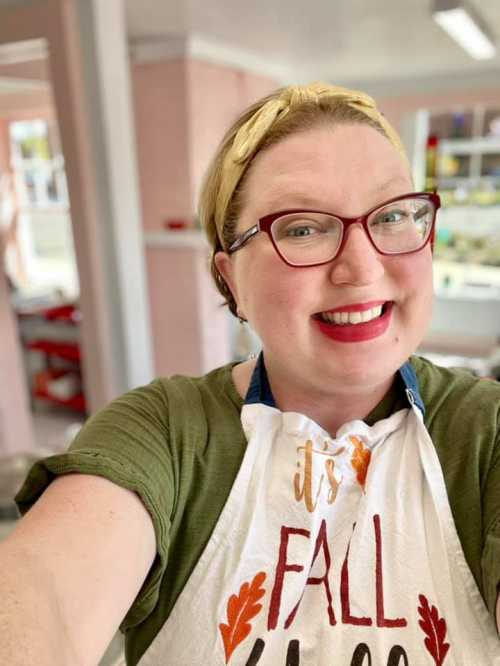 A smiling person with glasses and a headband, wearing a fall-themed apron, poses for a selfie in a bright, cheerful space.