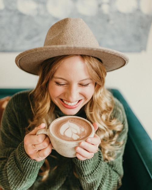 A smiling woman with long hair and a hat holds a cup of latte art, sitting in a cozy setting.