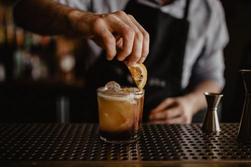 A bartender garnishes a cocktail with a lemon slice, ice visible in the glass on a metal bar surface.