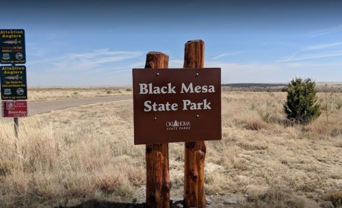 Sign for Black Mesa State Park, featuring wooden posts and a clear blue sky in the background.