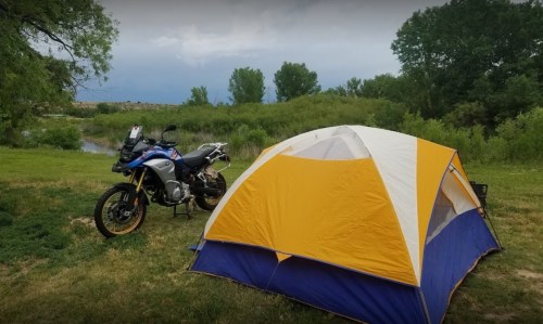 A blue and yellow tent set up beside a motorcycle in a grassy area near a river, with trees in the background.