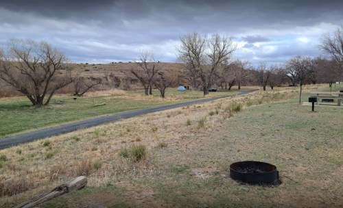 A grassy area with a path, trees, and two tents in the distance under a cloudy sky. A fire pit is in the foreground.