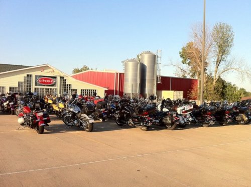A large gathering of motorcycles parked in front of a building with silos, under a clear blue sky.