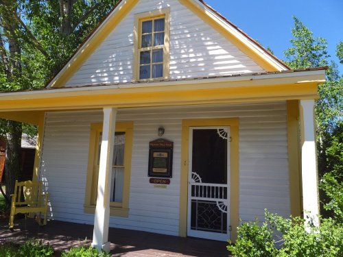 A charming white house with yellow trim, featuring a porch and an "Open" sign on the door. Lush greenery surrounds it.