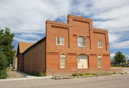 A brick building with a flat roof and windows, surrounded by greenery under a partly cloudy sky.