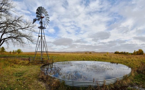 A windmill beside a circular water trough in a grassy landscape under a cloudy sky.