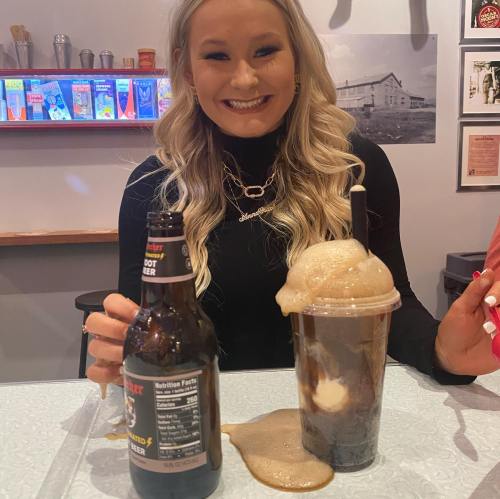 A smiling woman holds a frothy drink and a bottle, with a playful mess on the table in a lively setting.