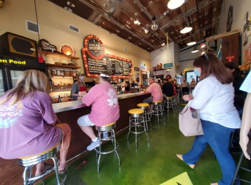 A lively restaurant interior with patrons sitting at a bar, colorful decor, and a menu board in the background.