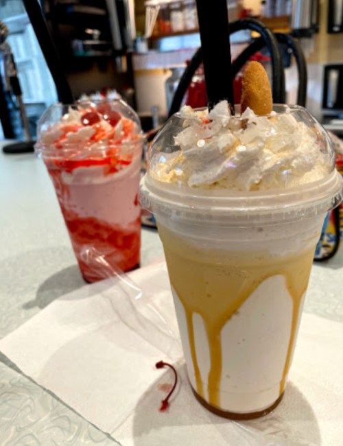 Two colorful milkshakes in clear cups, one topped with whipped cream and a cherry, the other with caramel drizzle.