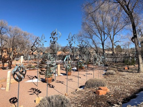 A garden featuring metal sculptures of plants and trees against a clear blue sky, surrounded by bare trees.