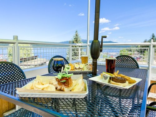 A scenic outdoor dining setup with plates of food and drinks, overlooking a lake and mountains under a clear blue sky.