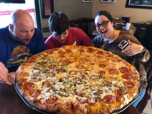Three people excitedly gaze at a giant pizza topped with pepperoni and vegetables, sitting at a restaurant table.