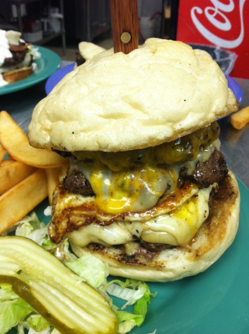 A large, stacked burger with melted cheese, served with fries, lettuce, and a pickle on a green plate.