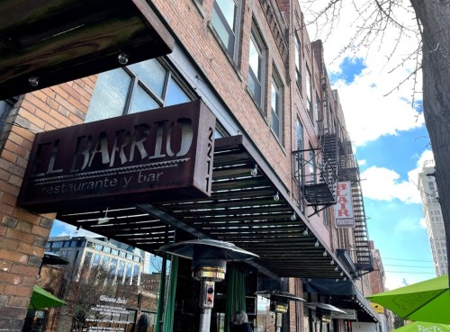 Sign for "El Barrio" restaurant and bar on a brick building, with a clear blue sky and cityscape in the background.