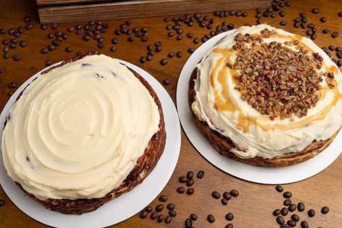 Two round cakes on white plates, one with smooth frosting and the other topped with nuts and caramel, surrounded by coffee beans.