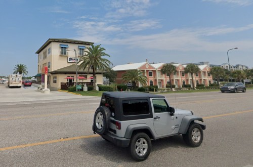 A silver Jeep drives past a restaurant and colorful buildings with palm trees on a sunny day.