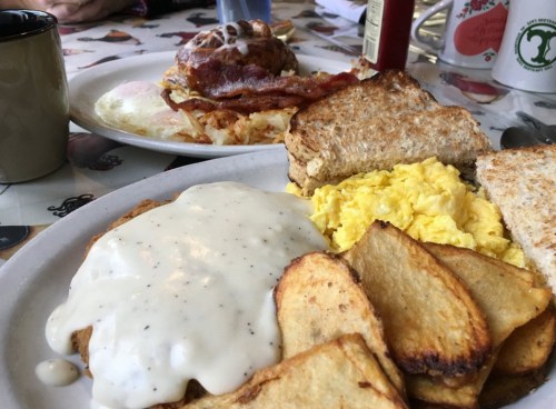 A plate of breakfast featuring scrambled eggs, biscuits with gravy, fried potatoes, and toast, with another dish in the background.
