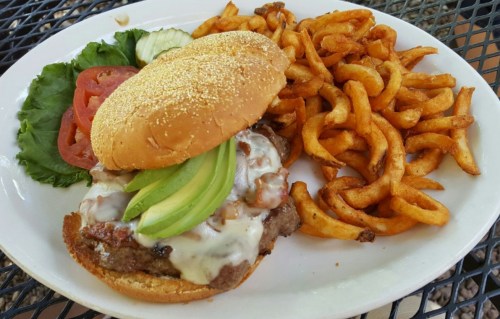A juicy burger with avocado, cheese, lettuce, and tomato, served with curly fries on a white plate.