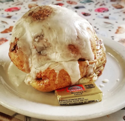 A large cinnamon roll topped with white icing, sitting on a plate next to a packet of butter.
