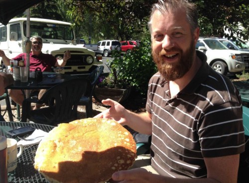 A man with a beard smiles while holding a large, round pancake at an outdoor table. A person sits in the background.