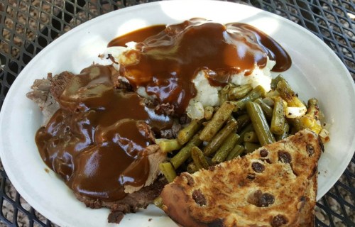 A plate of food featuring mashed potatoes with gravy, green beans, sliced beef, and a piece of bread.