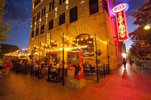 A lively outdoor dining area with string lights, people enjoying meals, and a neon sign on a historic building at dusk.