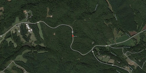 Aerial view of a winding road through dense forested area, with scattered fields and buildings nearby.