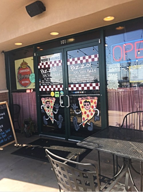 Exterior of a pizza restaurant with a "Now Hiring" sign, open door, and cartoon pizza characters on the windows.