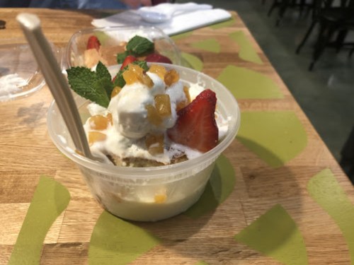 A dessert in a clear cup with whipped cream, strawberries, and mint, placed on a wooden table.