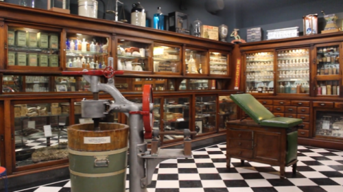 A vintage apothecary with wooden cabinets, glass jars, and a green examination chair on a black and white checkered floor.