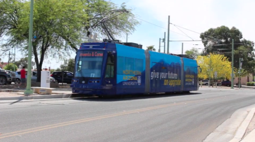A blue tram with university branding travels along a street lined with trees and parked cars.