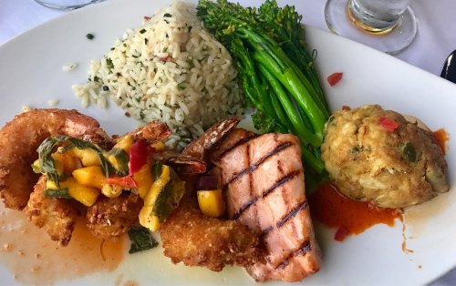 A plate featuring grilled salmon, shrimp with mango salsa, rice, broccolini, and a seafood stuffing ball.