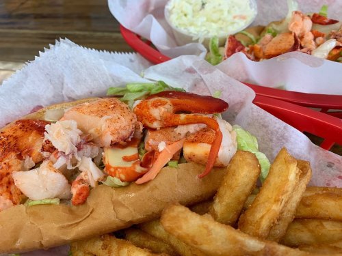 A lobster roll with lettuce on a bun, served with golden fries and a side of coleslaw in a red basket.