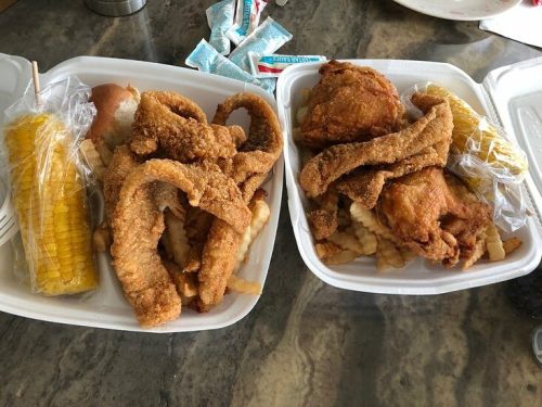 Two takeout containers with fried chicken, crinkle-cut fries, fried fish, and corn on the cob, accompanied by condiments.