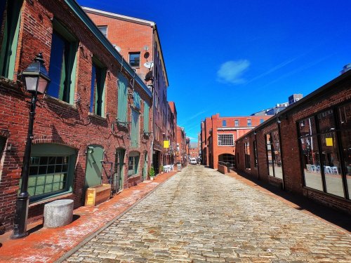 A sunny cobblestone street lined with red brick buildings and large windows, creating a charming urban atmosphere.