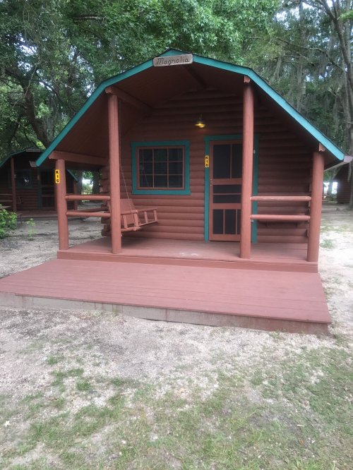 A rustic wooden cabin with a porch, swing, and green accents, surrounded by trees and gravel.