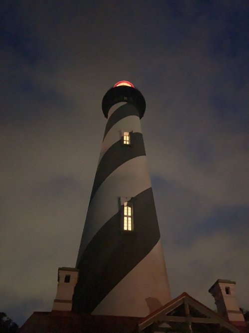 A tall striped lighthouse illuminated at night, with a glowing red light at the top and a cloudy sky in the background.