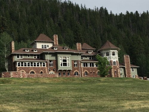 A large, multi-story house with a mix of stone and wood, surrounded by trees and a grassy area.