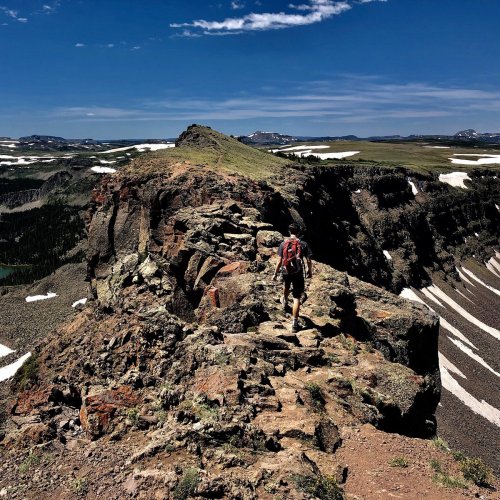 A hiker walks along a rocky ridge with expansive mountain views and blue skies in the background.