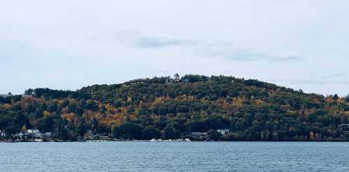 A scenic view of a lake with a forested hill in the background, featuring autumn foliage and a cloudy sky.